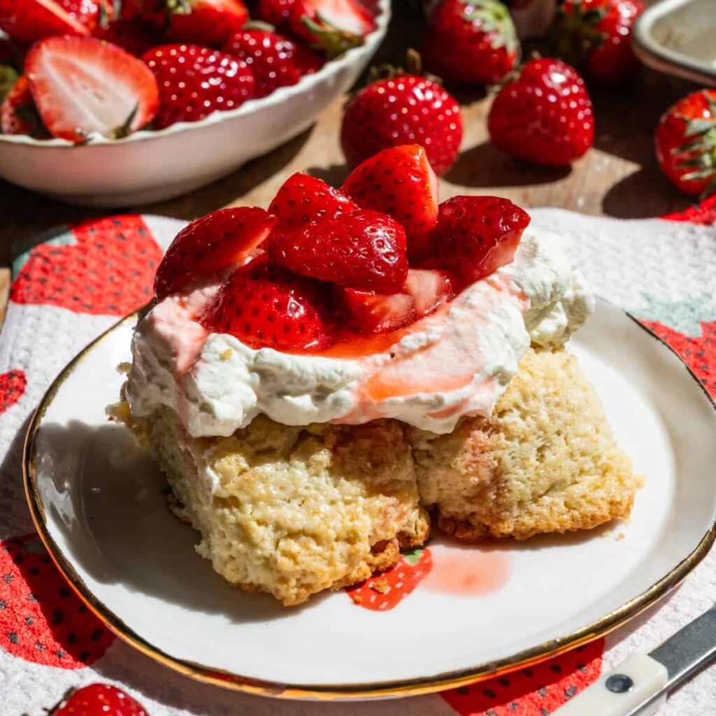 Slice of strawberry shortcake on a plate with strawberries in the background.