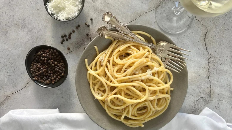 Bol gris avec des pâtes cacio e pepe dedans, Ramekin de grains de poivre noir, Ramekin de parmesan frais râpé et verre de vin blanc