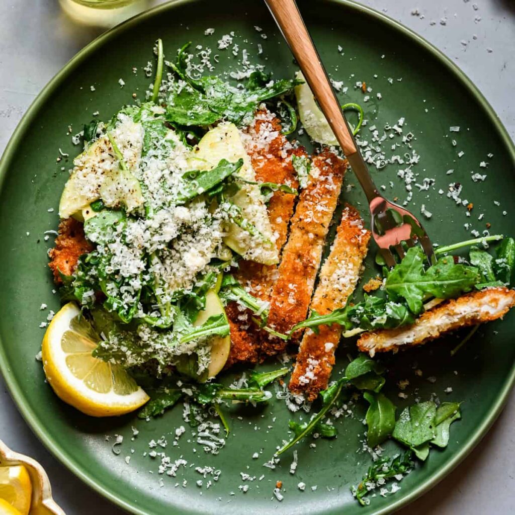 Overhead view of chicken cutlet topped with arugula and shaved apple salad and grated parmesan.