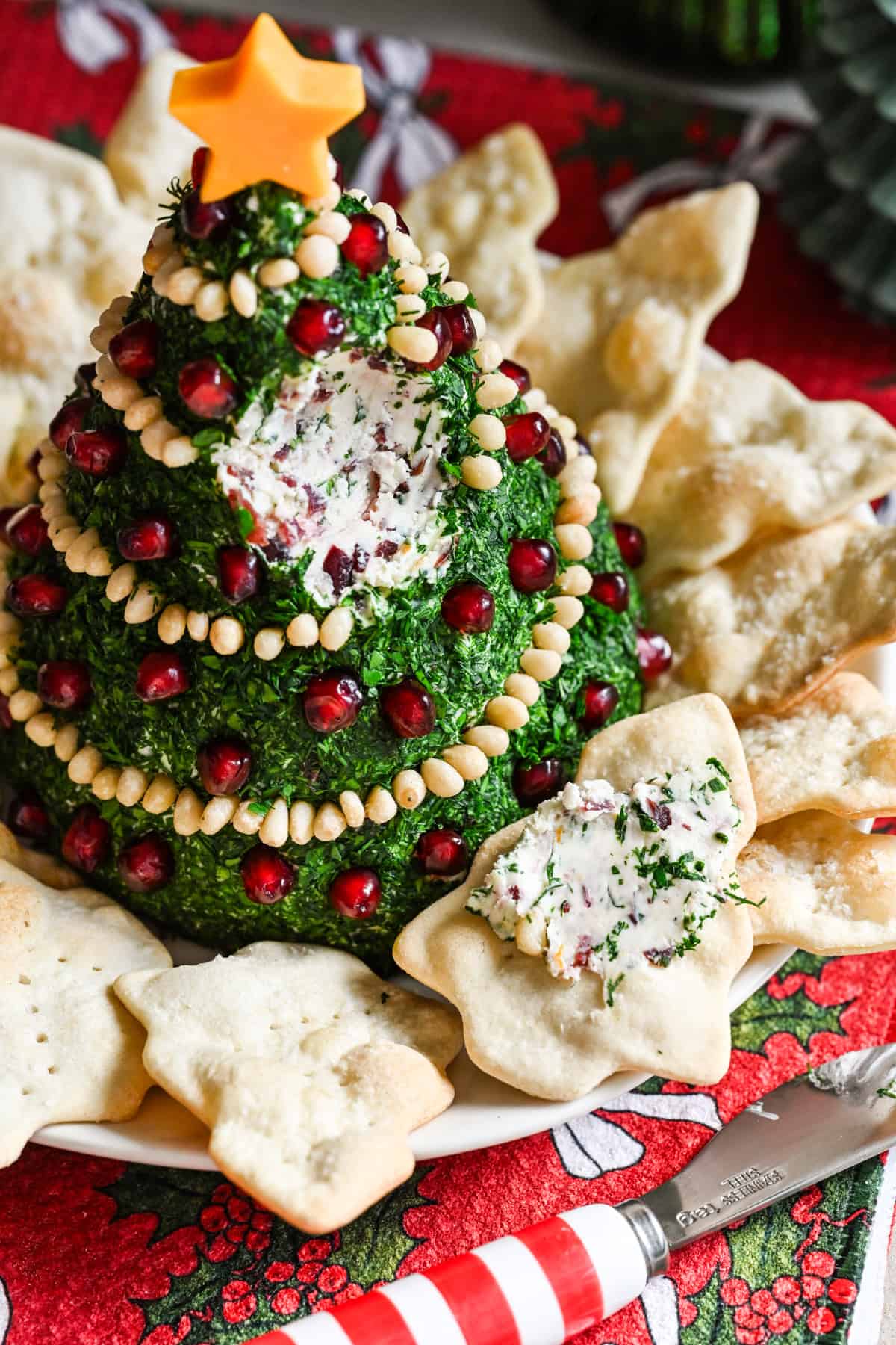 Boule de fromage en forme d'arbre de Noël recouverte d'herbes et décorée de guirlandes de pignons de pin et d'ornements de grenades.