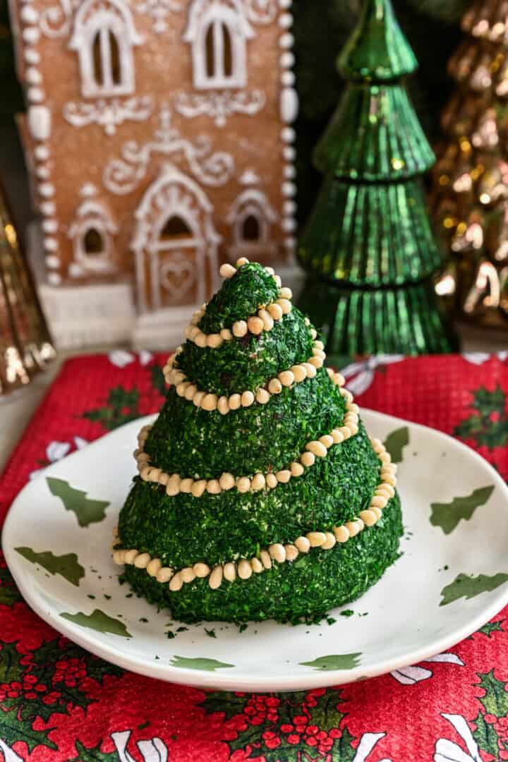 Boule de fromage en forme de sapin de Noël enrobée d'herbes hachées et décorée d'une guirlande de pignons de pin.