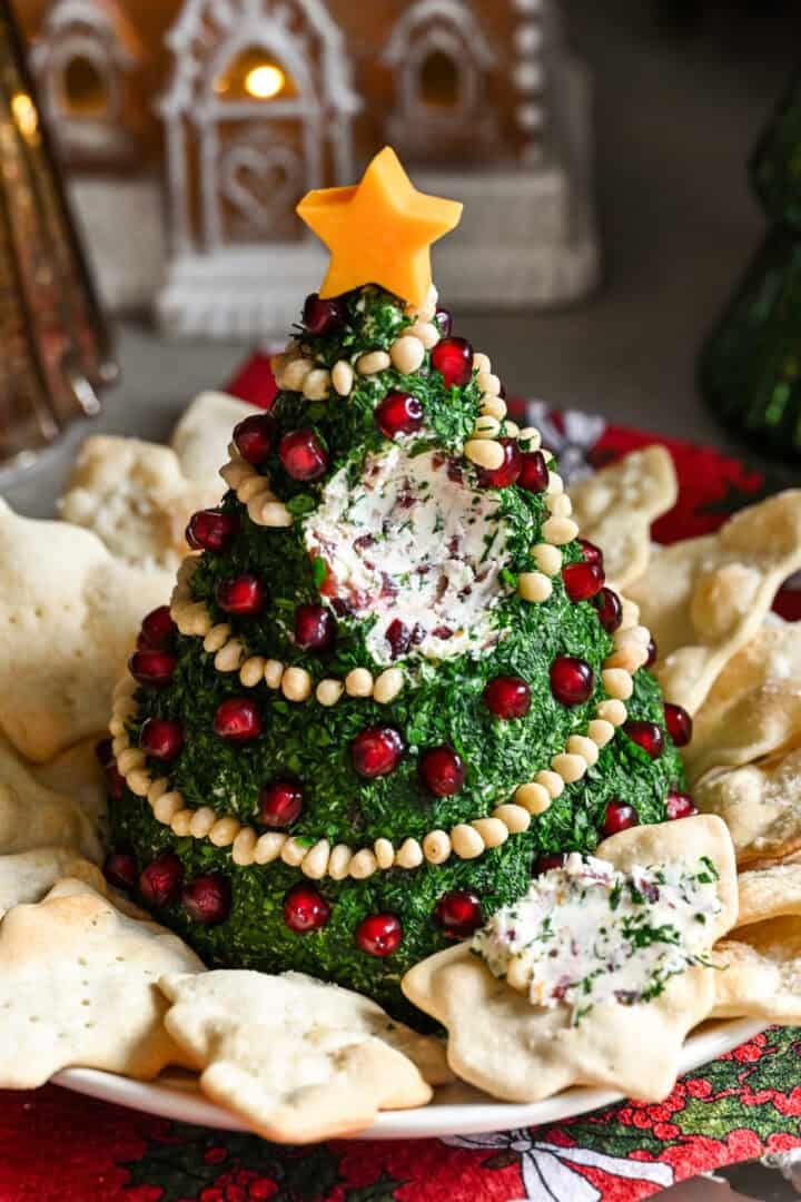 Boule de fromage en forme d'arbre de Noël recouverte d'herbes et décorée de guirlandes de pignons de pin et d'ornements de grenades.