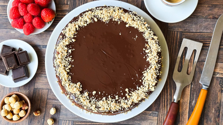 Un gâteau au chocolat sans farine accompagné de framboises, de carrés de chocolat, de noisettes grillées et d'ustensiles de cuisine.