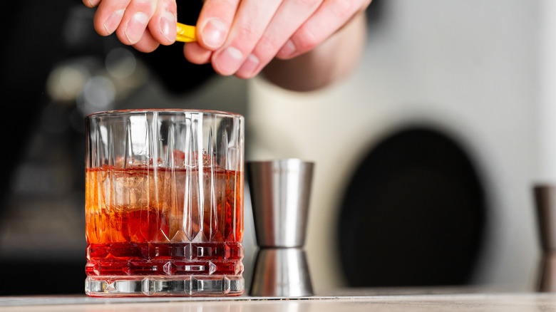 On voit les mains d'une personne pressant un zeste d'orange sur un cocktail dans un verre à whisky.