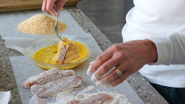 Mains draguant le poulet cru dans la farine, l'œuf et la chapelure sur une longue feuille de papier sulfurisé
