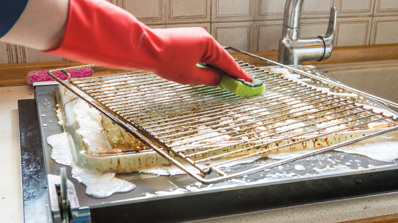 On voit la main gantée d’une personne frottant une grille de four au-dessus d’un évier de cuisine.