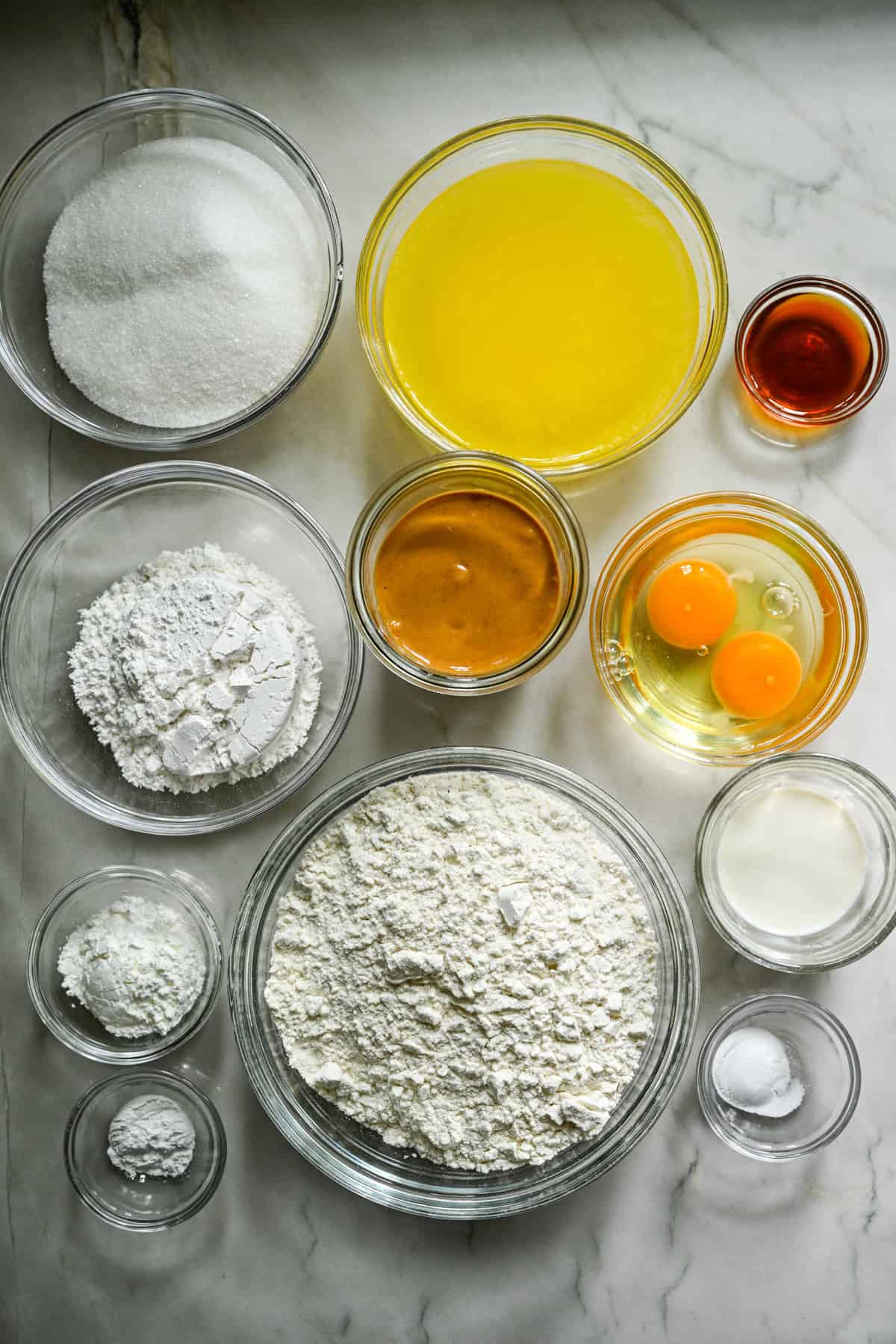 Vue aérienne des ingrédients pour les beurres de noix faits maison dans des bols de préparation séparés sur un comptoir blanc.