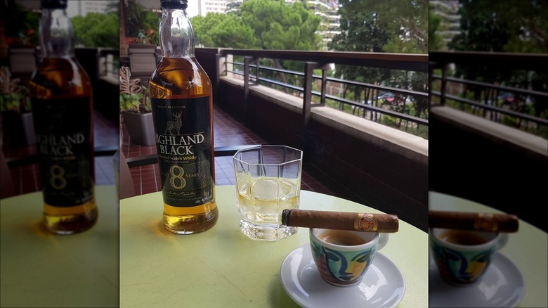 Une bouteille de scotch Highland Black d'Aldi sur la table du balcon d'un appartement à côté d'un verre à whisky rempli de glace et de whisky, et un cigare en équilibre autour d'une tasse de café