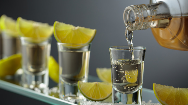 Tequila étant versée d'une bouteille dans une rangée de verres à shot avec des citrons verts.