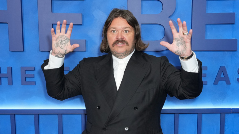 L'acteur Matty Matheson pose les mains en l'air devant un mur bleu.