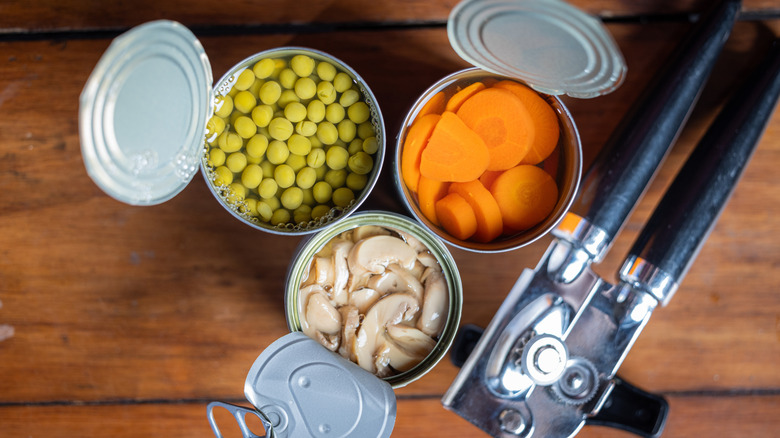 Boîtes ouvertes de pois, de carottes et de champignons sur une table en bois avec un ouvre-boîte