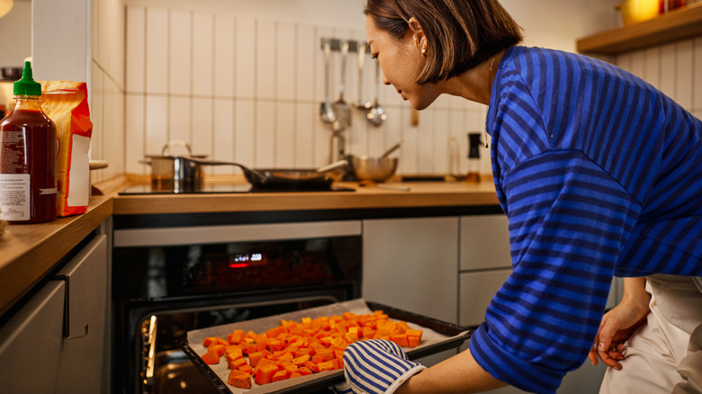 Une femme place une plaque recouverte de papier sulfurisé et remplie de cubes de courge dans le four dans une cuisine moderne
