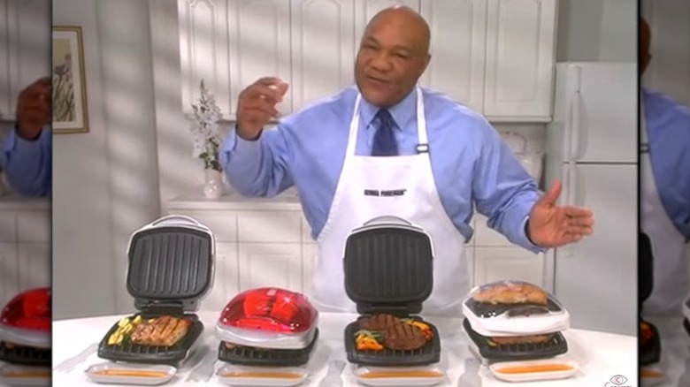 George Foreman portant un tablier debout derrière quatre grilles George Foreman dans un publi-reportage