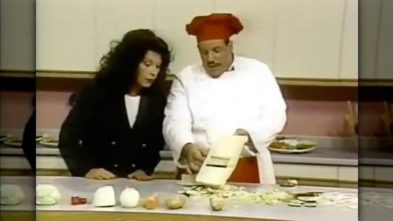 Un homme vêtu d'une blouse de chef et d'une toque rouge tranche un oignon sur un Kitchen Wizard Plus tandis qu'une femme vêtue d'un blazer noir regarde