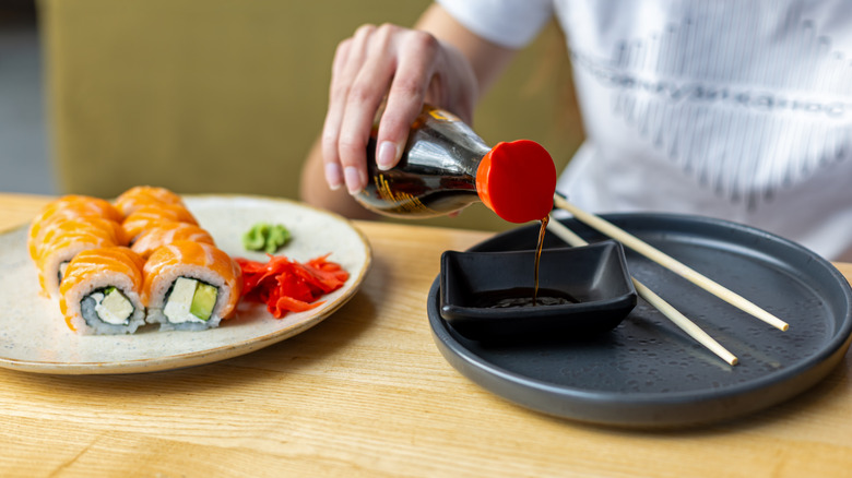 Gros plan de la sauce soja versée dans un bol à tremper à côté des rouleaux de sushi au saumon, des baguettes, du gingembre et du wasabi.