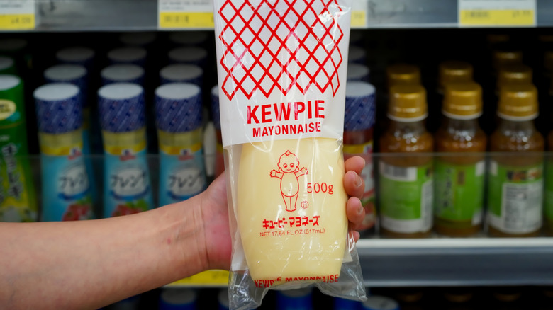 Gros plan de la mayonnaise de marque Kewpie de style japonais tenue en main dans une allée de supermarché local avec des étiquettes de produits visibles en arrière-plan.