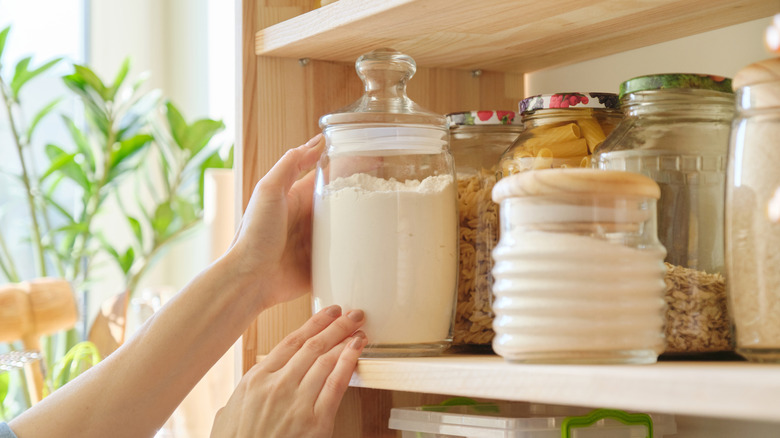 On voit la main d’une personne saisissant un pot en verre de farine sur une étagère.