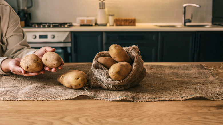 Mains tenant et disposant des pommes de terre fraîches dans un sac en toile de jute sur une table en bois dans une cuisine confortable. Éclairage chaleureux et ambiance rustique.