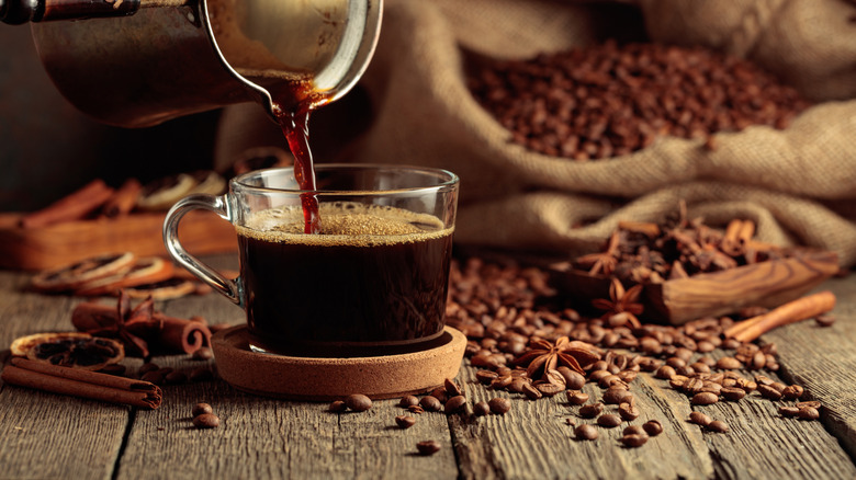 Une tasse de café fraîchement moulue est servie dans une cafetière avec des grains de café, de la cannelle et d'autres épices sur une table autour de la tasse