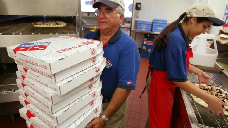 Deux employés Domino's dans la cuisine d'un magasin Domino's, l'un portant des boîtes de pizza et l'autre préparant une pizza prête à être mise au four.
