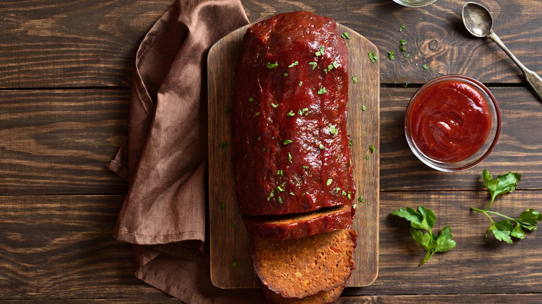 Pain de viande sur une planche de bois, avec des herbes, un bol de sauce et un torchon sur le côté