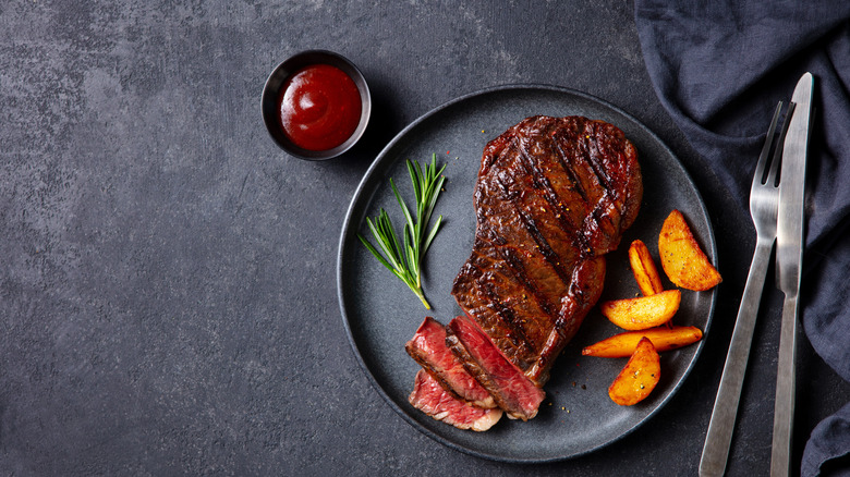Une assiette de steak cuit avec des pommes de terre et un brin de romarin frais, à côté de couverts et d'un ramequin de sauce