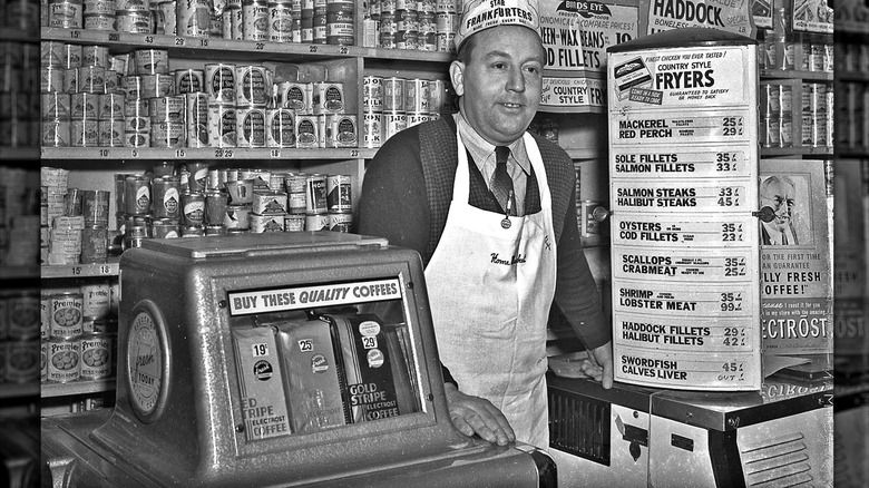 Photo en noir et blanc d’un homme debout au comptoir d’une épicerie devant une machine à café en grains et une liste de prix pour les fruits de mer.