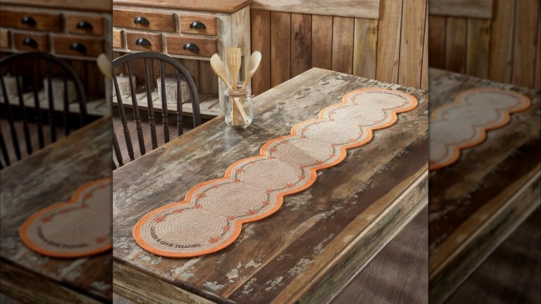 Un chemin de table tissé avec de petites citrouilles qui le bordent, posé sur une table en bois avec un pot d'ustensiles de cuisine à proximité.