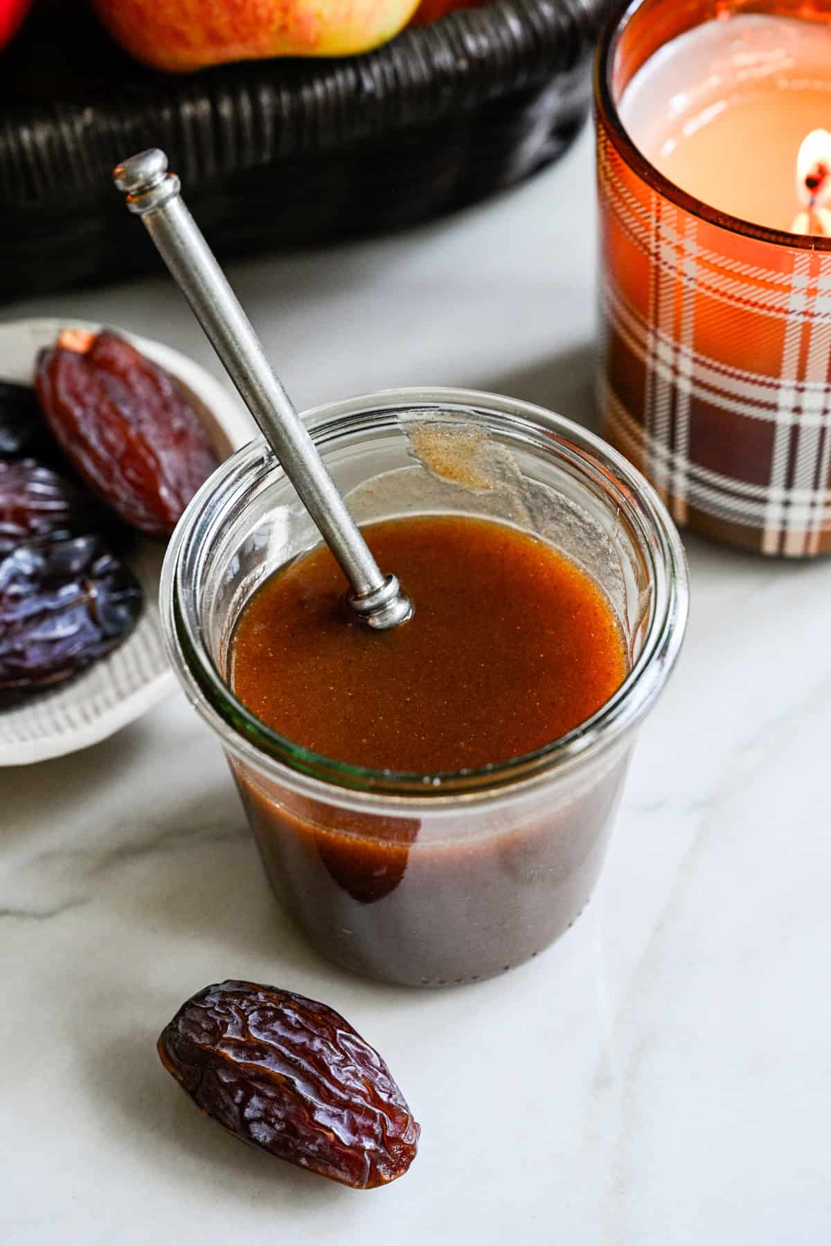 Sauce caramel aux dattes dans un bocal en verre avec une bougie d'automne et un bol de dattes en arrière-plan.