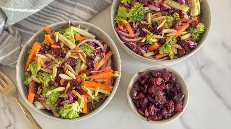 deux bols de salade de brocoli sucrée et salée à côté d'un bol de canneberges séchées