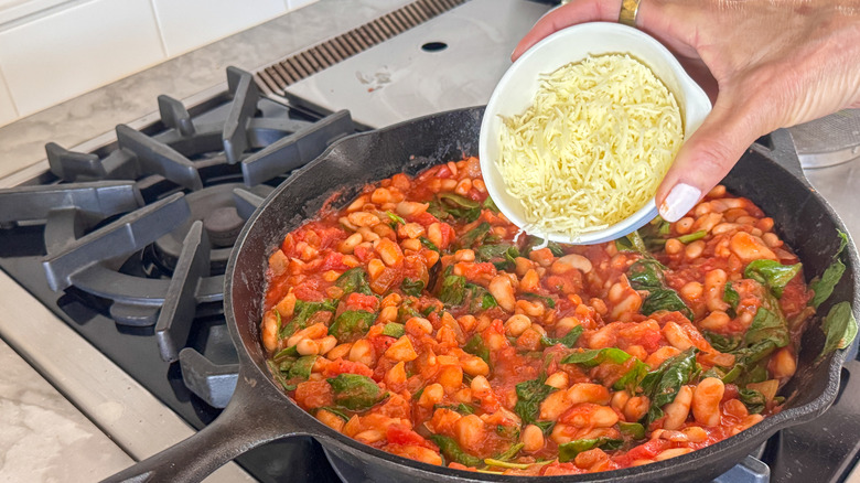 Verser à la main un bol de fromage râpé dans une poêle avec des haricots, de la sauce et des épinards