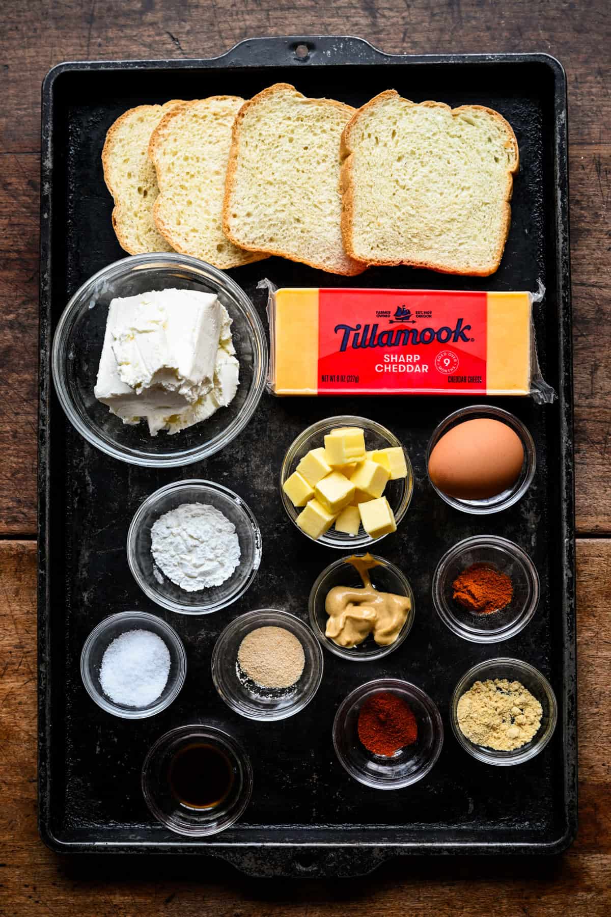 Vue aérienne des ingrédients des rêves de fromage sur une plaque sombre : du pain, du fromage à la crème, un bloc de cheddar fort Tillamook, du beurre en cubes, un œuf, de la moutarde et des petits bols d'épices.