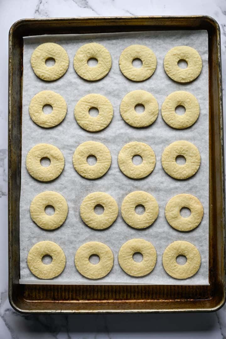 Vingt biscuits sablés en forme d'anneau, doré pâle avec un motif en relief, sur une plaque à pâtisserie tapissée de papier sulfurisé.