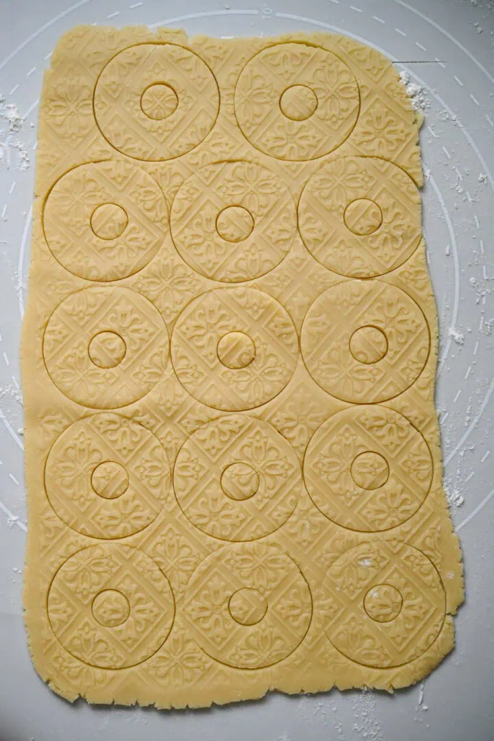 Pâte sablée étalée, gaufrée d'un motif de carreaux floraux et découpée en rondelles avec de petits trous centraux, avant de retirer les biscuits.