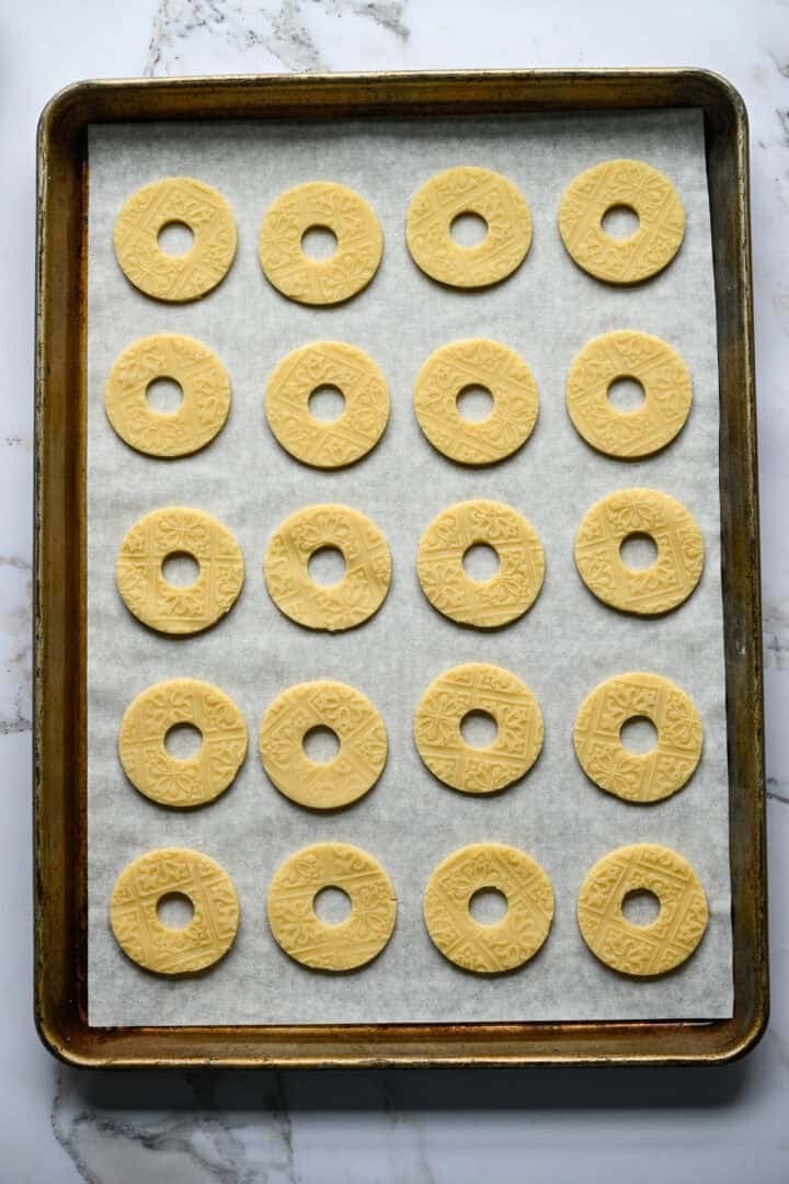 Vingt biscuits sablés non cuits en forme d'anneau avec un motif en relief alignés en rangées sur une plaque à pâtisserie tapissée de papier sulfurisé.
