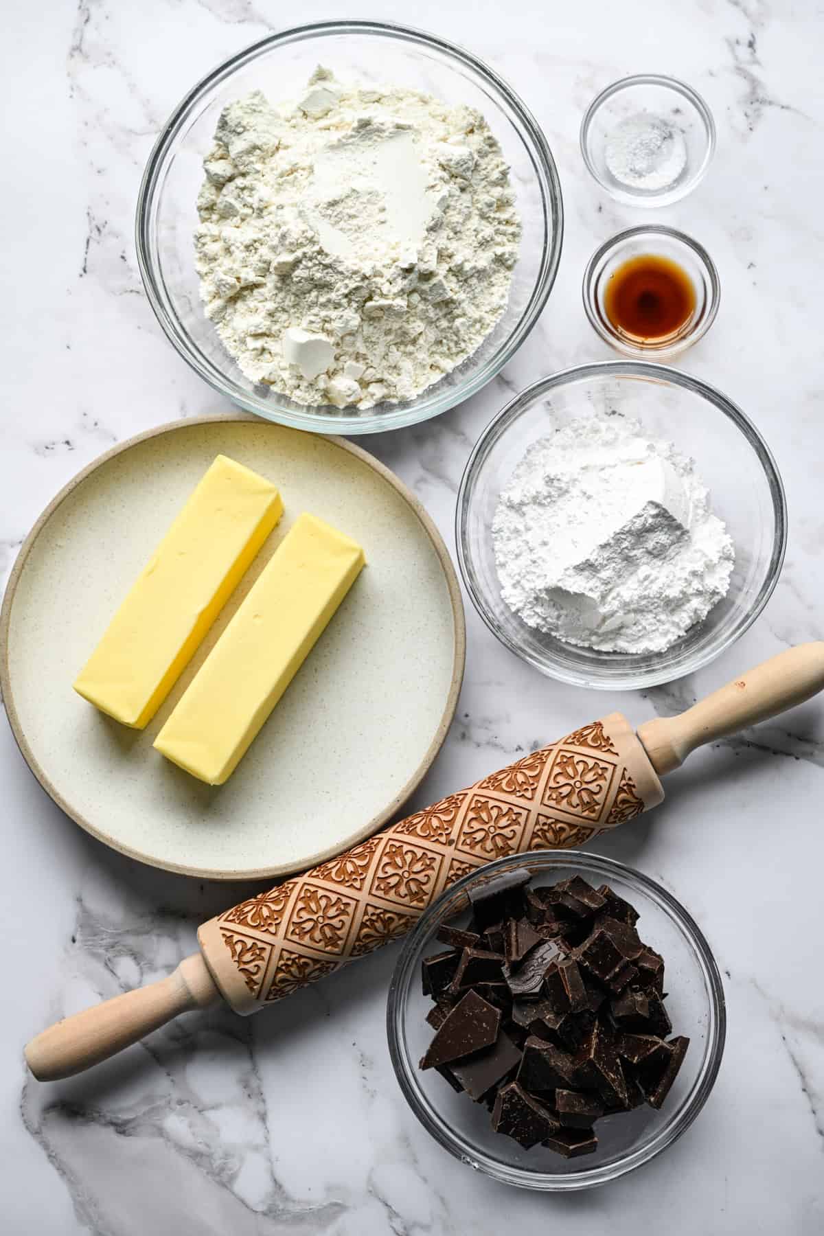 Bols en verre de farine, sucre en poudre, chocolat noir haché, vanille et sel sur un comptoir en marbre avec deux bâtons de beurre sur une assiette et un rouleau à pâtisserie en bois gaufré.