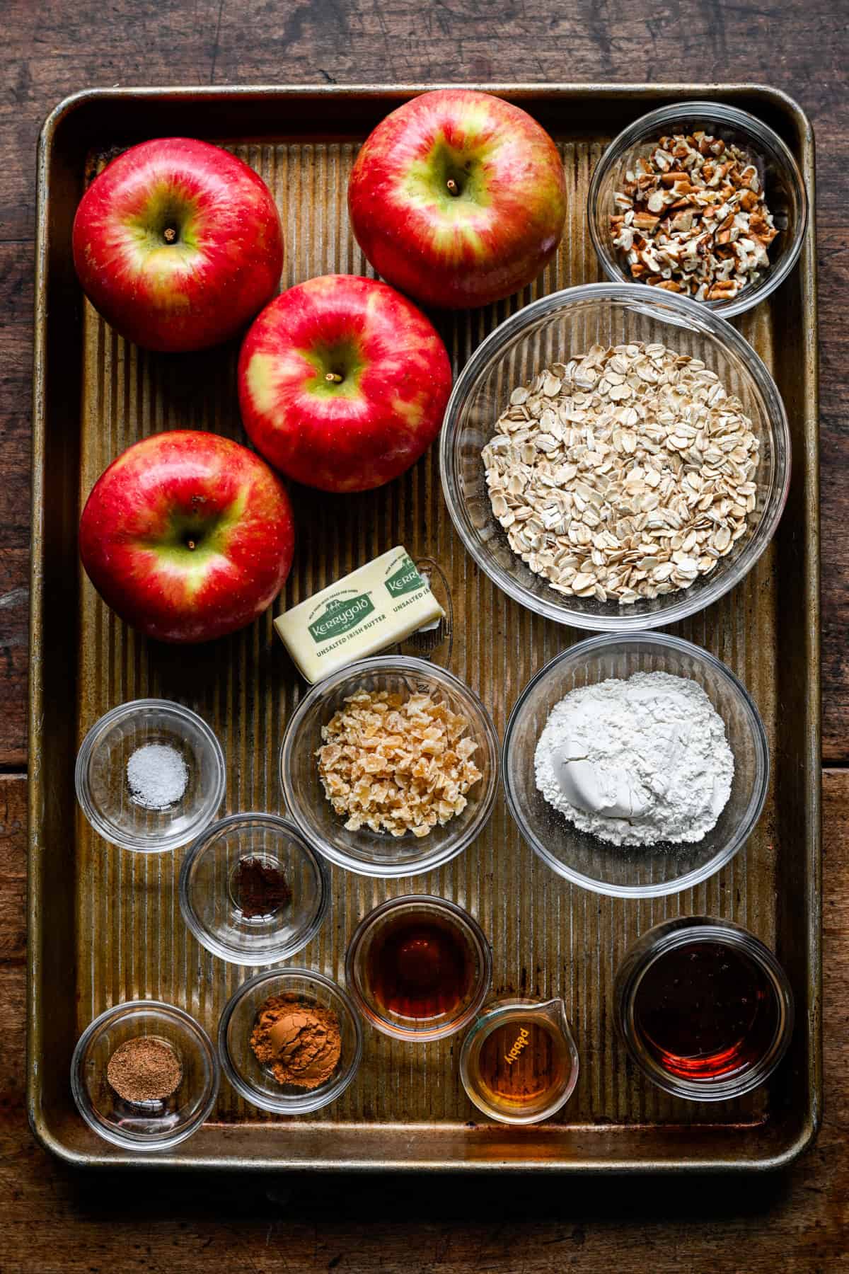 Ingrédients pour pommes au four sur une plaque à pâtisserie : quatre pommes Honeycrisp, des bols de flocons d'avoine, des pacanes hachées, de la farine, du gingembre confit, un bâton de beurre, des épices moulues, du sirop d'érable et de la vanille.