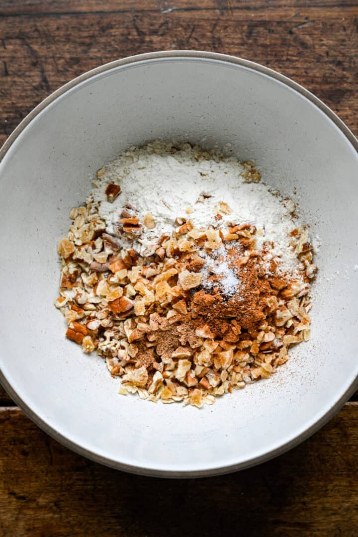 Flocons d'avoine, pacanes hachées, gingembre confit, farine, cannelle, muscade, clous de girofle et sel dans un bol à mélanger blanc avant de remuer.