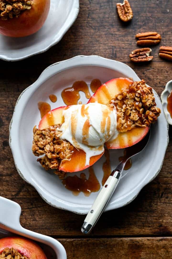 Une pomme au four coupée en deux avec de la glace à la vanille et du caramel aux dattes, une deuxième portion et des pacanes éparpillées en arrière-plan.