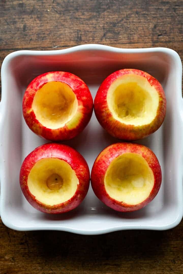 Quatre pommes Honeycrisp, évidées avec le fond intact, placées dans un plat carré blanc allant au four.
