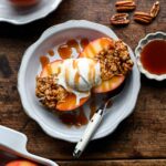A baked apple cut in half on a plate with a scoop of vanilla ice cream, drizzled with date caramel, pecans and a small dish of the caramel alongside.