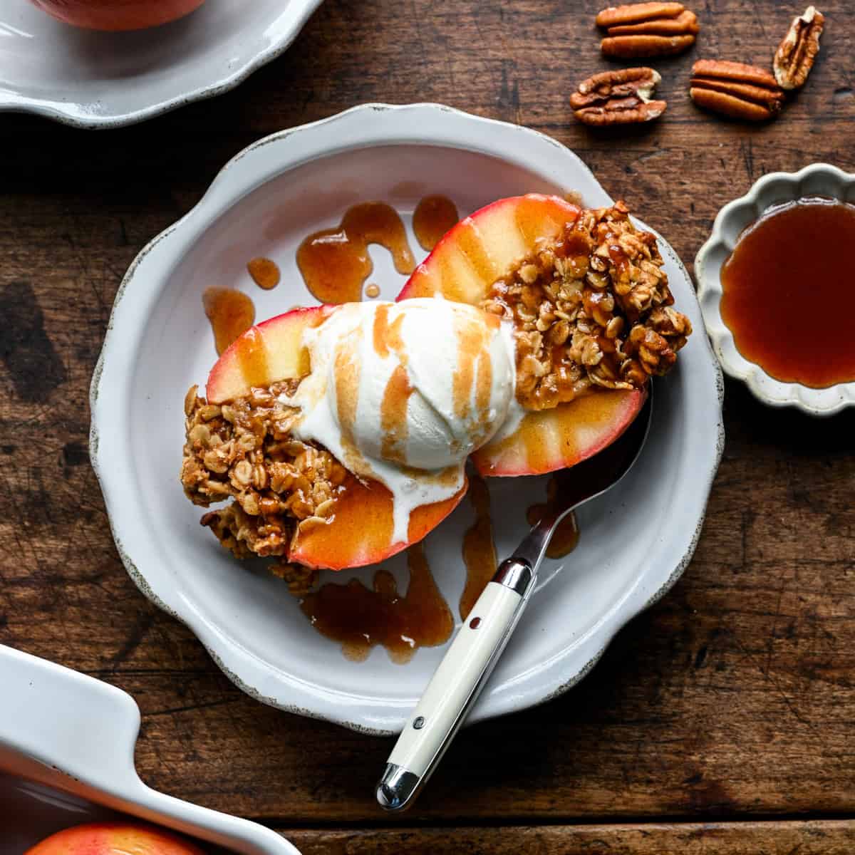 Une pomme au four coupée en deux sur une assiette avec une boule de glace vanille, arrosée de caramel aux dattes, des noix de pécan et un petit plat de caramel à côté.