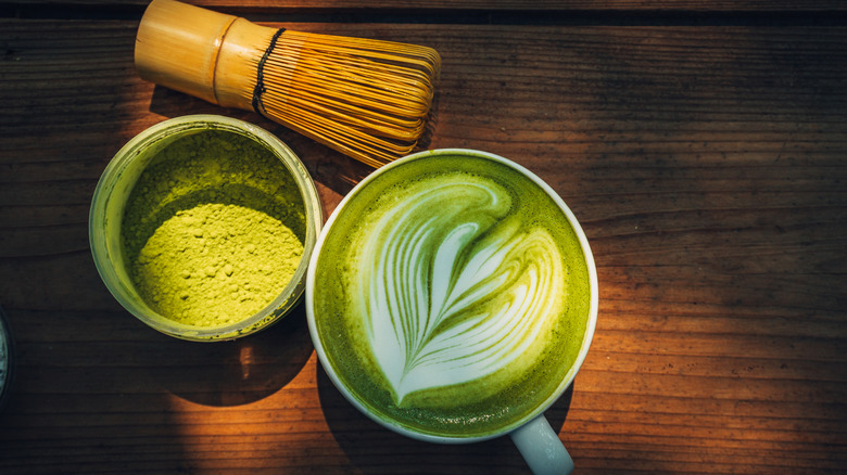 Un matcha latte à côté d'un pot de poudre de matcha et d'un Chasen