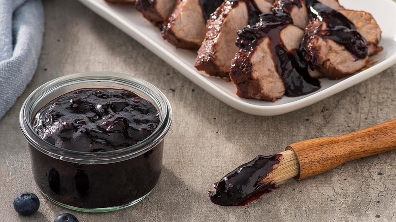 Un pot de sauce barbecue aux bleuets à côté d'un pinceau et d'une assiette de tranches de porc