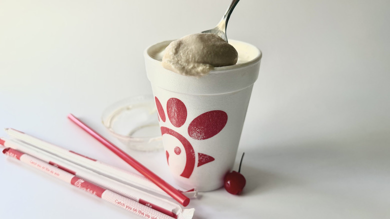 Milkshake au chocolat Chick-fil-A avec une cuillère insérée dans la tasse à côté d'une cerise, des pailles et du couvercle