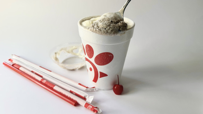 Chick-fil-A's Cookies & Cream Milkshake avec une cuillère insérée dans la tasse à côté d'une cerise, des pailles et du couvercle