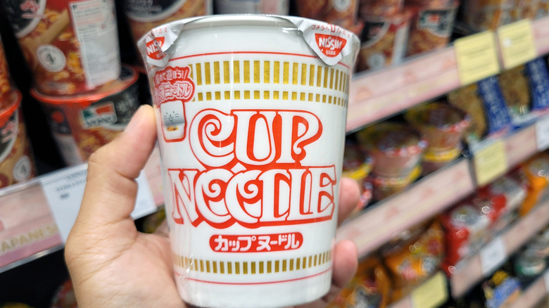 Une main tenant une Nissin Cup Noodle dans un supermarché, le long de l'allée des ramen instantanés.