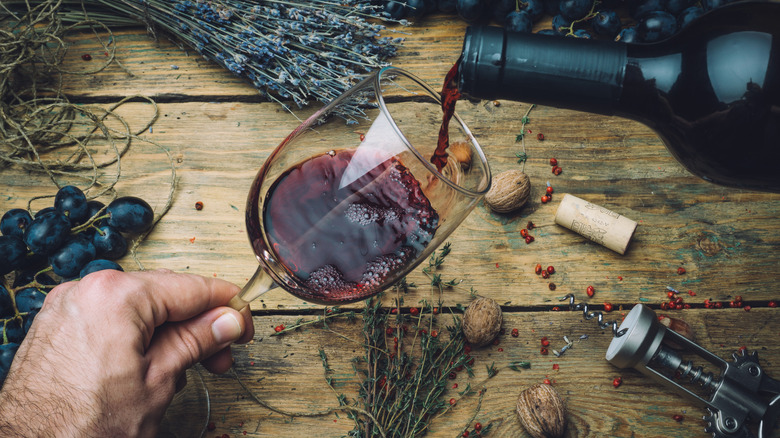 Un individu versant du vin rouge à déguster devant des raisins, des herbes et un tire-bouchon