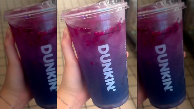 Main tenant une limonade Starlight, avec une couche supérieure violette et une couche inférieure indigo avec des morceaux de fruit du dragon, dans une tasse Dunkin' en plastique transparent