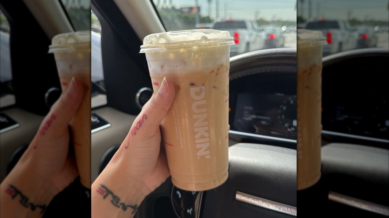 Main de femme avec des tatouages ​​tenant un café au lait glacé dans une tasse Dunkin en plastique transparent devant un volant